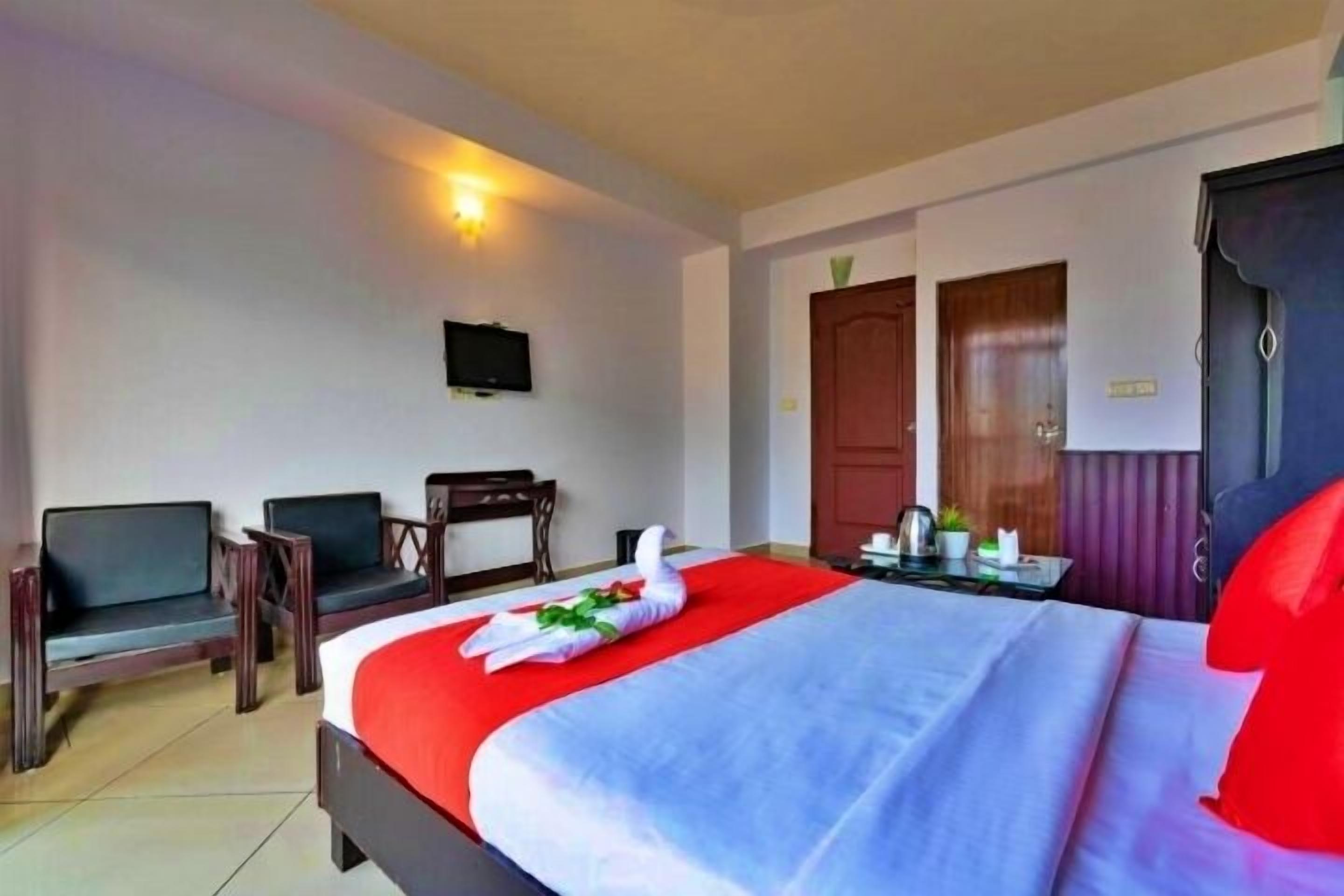 A hotel room with a bed, two chairs, a TV, and a desk with a kettle.
