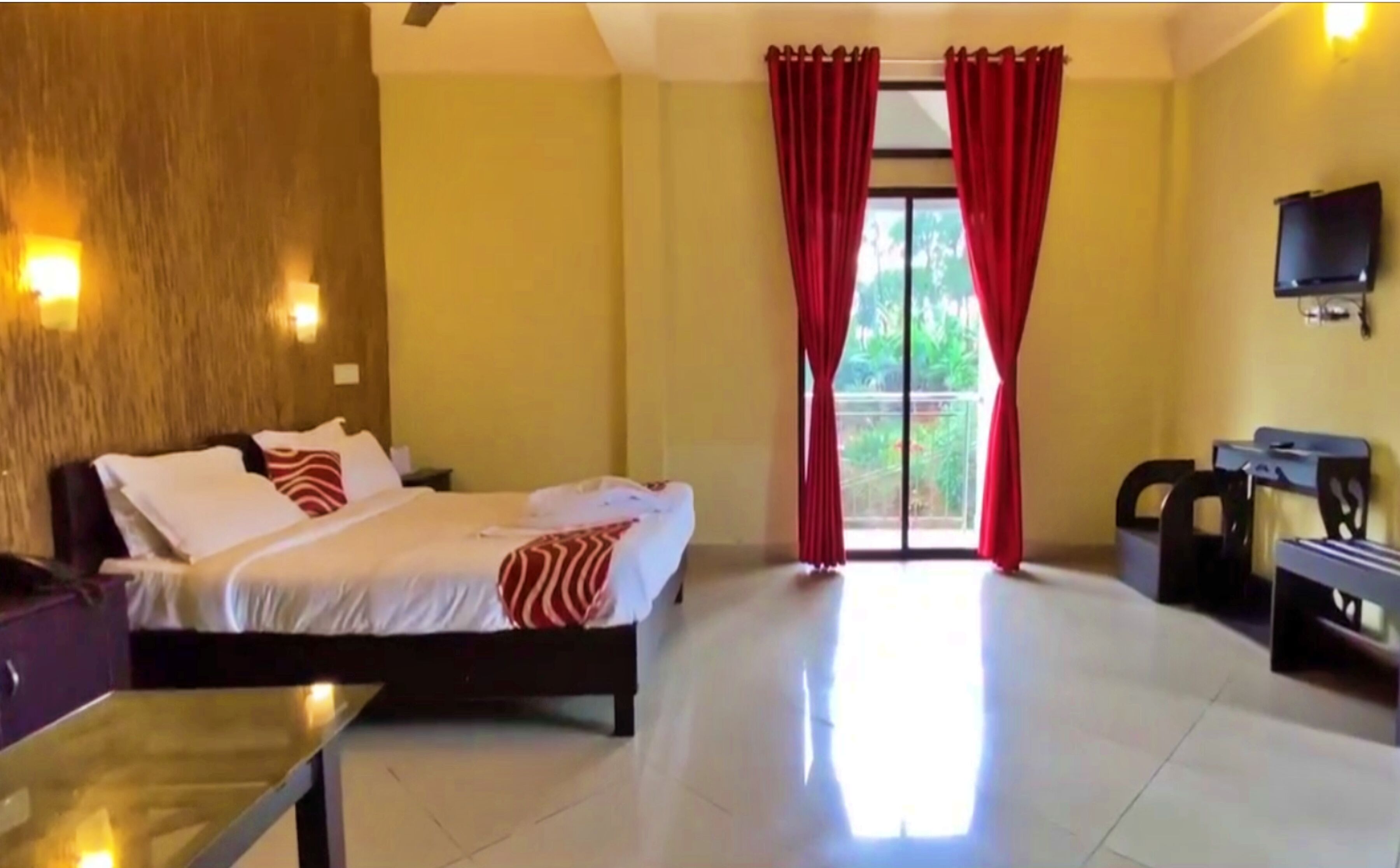 A hotel room with a bed, a desk, a chair, a TV, and a balcony with red curtains.