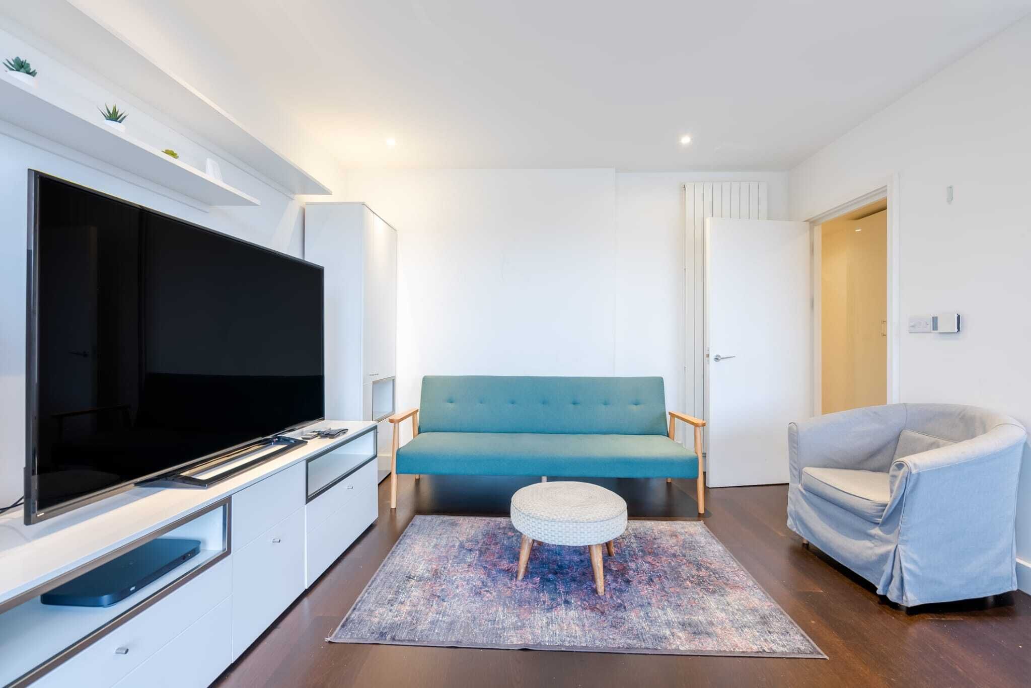 Uma sala de estar moderna com televisão de tela plana, sofá verde-água, otomano redondo e poltrona cinza claro.