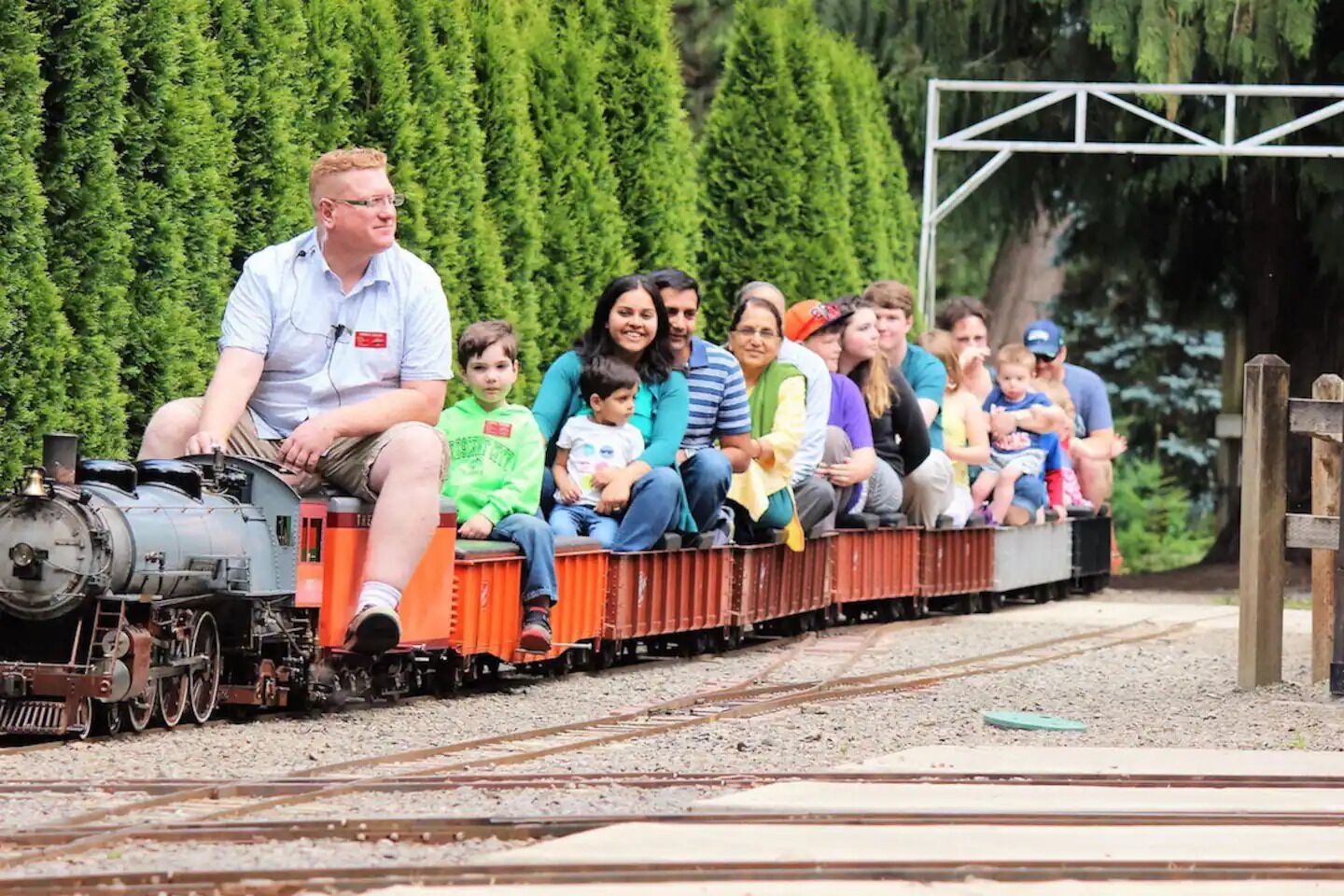 A miniature train with passengers, set on a track with a backdrop of green hedges.