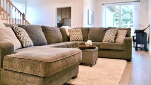 Ein modernes Wohnzimmer mit Ecksofa, Hocker und einem großen Fenster, das für natürliches Licht sorgt.