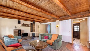 Amplia sala de estar con vigas de madera en el techo, chimenea, televisor y un rincón de descanso acogedor con dos sillones y un sofá.