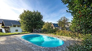 Una piscina con borde de piedra, rodeada de vegetación y casas.