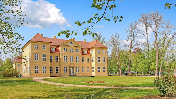 Un grand bâtiment jaune, surmonté d’un toit rouge, entouré d’une pelouse bien entretenue et d’arbres.