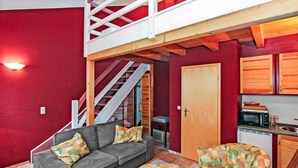 Un salon chaleureux avec un canapé gris et un fauteuil à motifs, un escalier en bois et un petit espace cuisine.