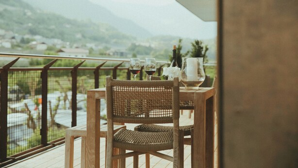 En balkon med et bord dækket til to personer, komplet med en vinflaske, to vinglas og et stearinlys. En fletstol er placeret ved bordet.