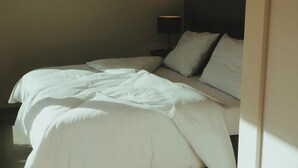 En pænt redt seng med hvide sengetøj og puder, en sengebordlampe og en mørk sengegavl.