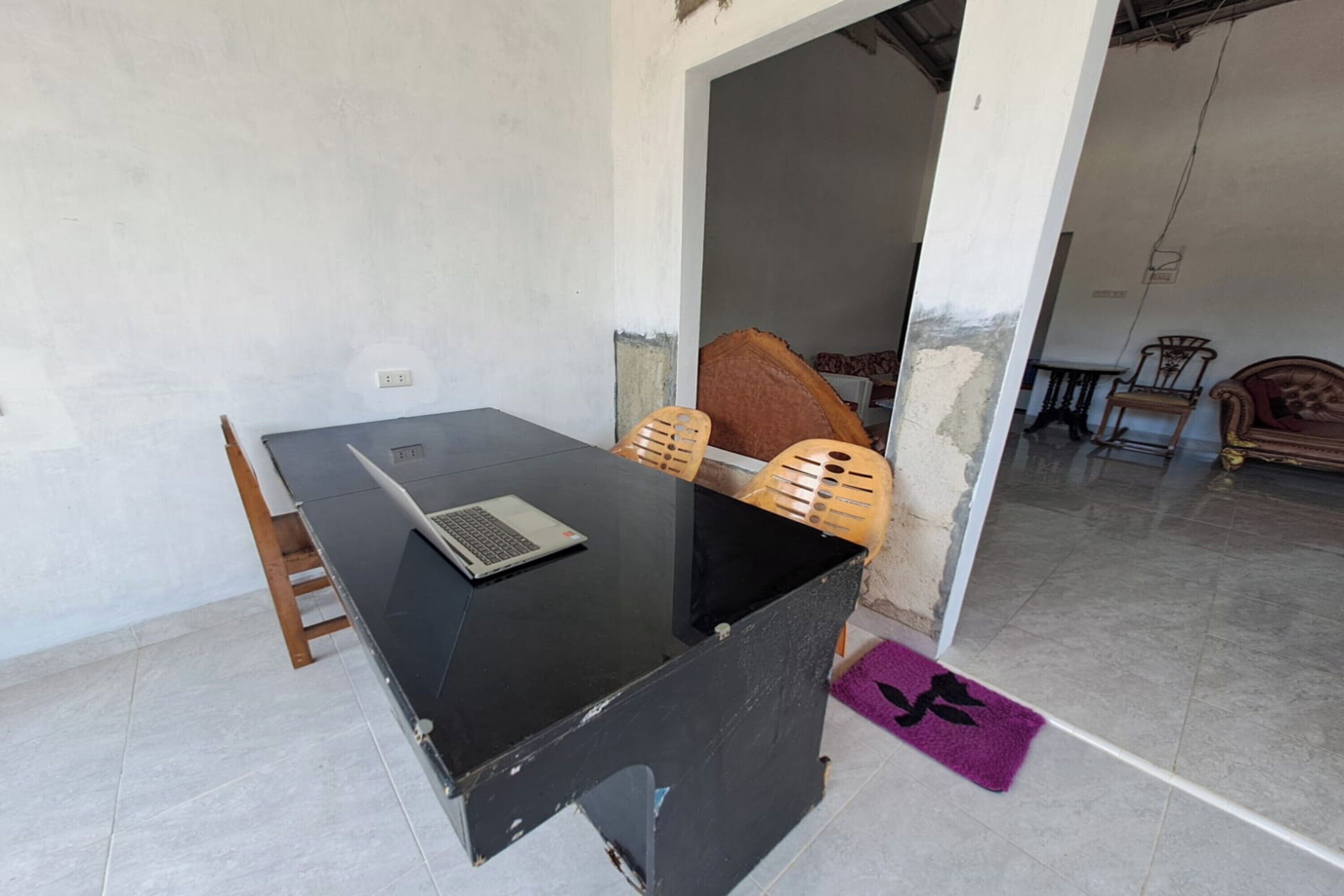A room with a black desk, a laptop, and a chair. There is a doorway leading to another room with a bed and a chair.