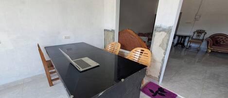 A room with a black desk, a laptop, and a chair. There is a doorway leading to another room with a bed and a chair.