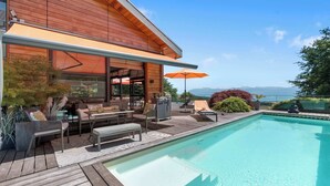 Una casa moderna con fachada de madera, piscina, mobiliario exterior y vistas a las montañas.