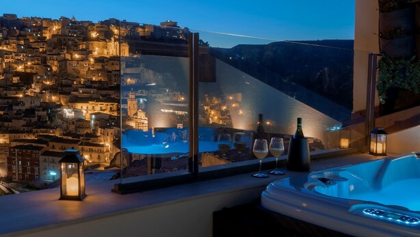 A balcony with a hot tub, two wine glasses, and a bottle. A cityscape with buildings and a river is visible through a glass partition. - Dimora Bonafede Ibla Design Rooms (Ragusa)