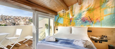A hotel room with a bed, a wooden ceiling, a colorful abstract painting, a balcony with a view of a cityscape, and a small table with a phone.
