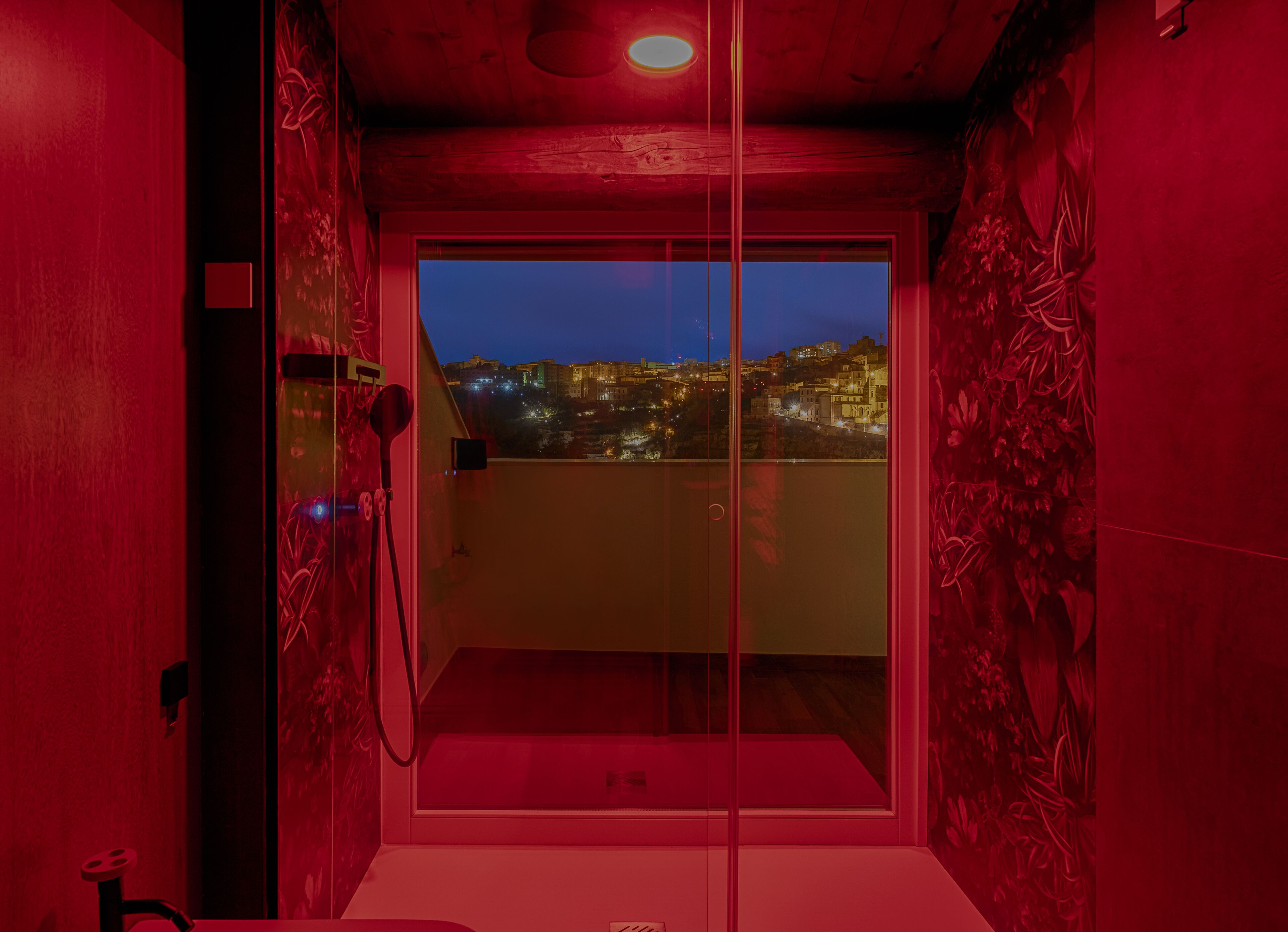 A room with a glass door leading to a balcony with a city view at dusk.