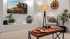 Een moderne woonkamer met een grote televisie waarop 'PALM SPRINGS' staat, een houten tafel met een charcuterieplank, een lamp en decoratieve objecten.