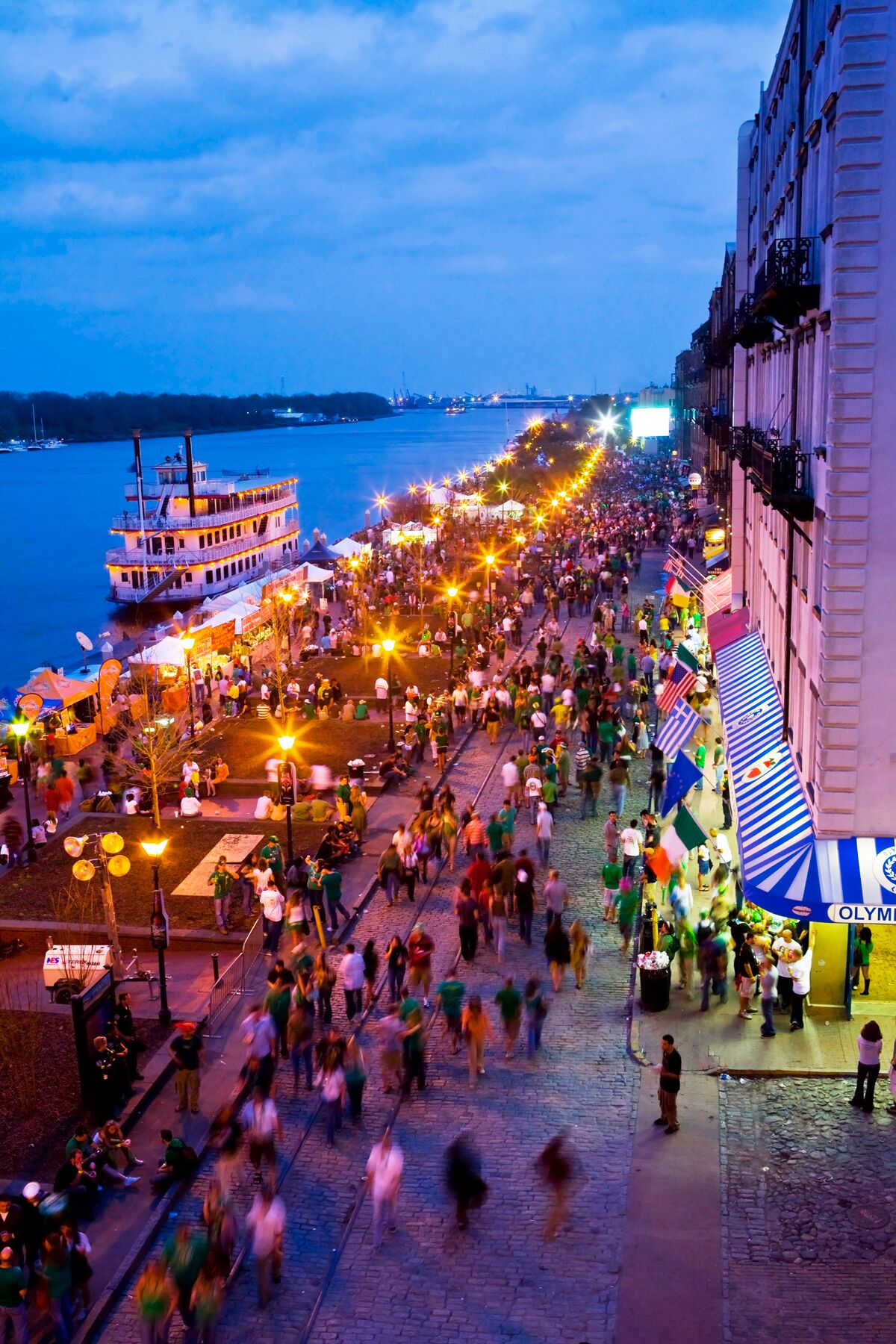 Eine Uferpromenade mit einem am linken Ufer vertäuten Boot, einer Menschenmenge und einem Gebäude mit blauen und weißen Streifen an der rechten Seite.