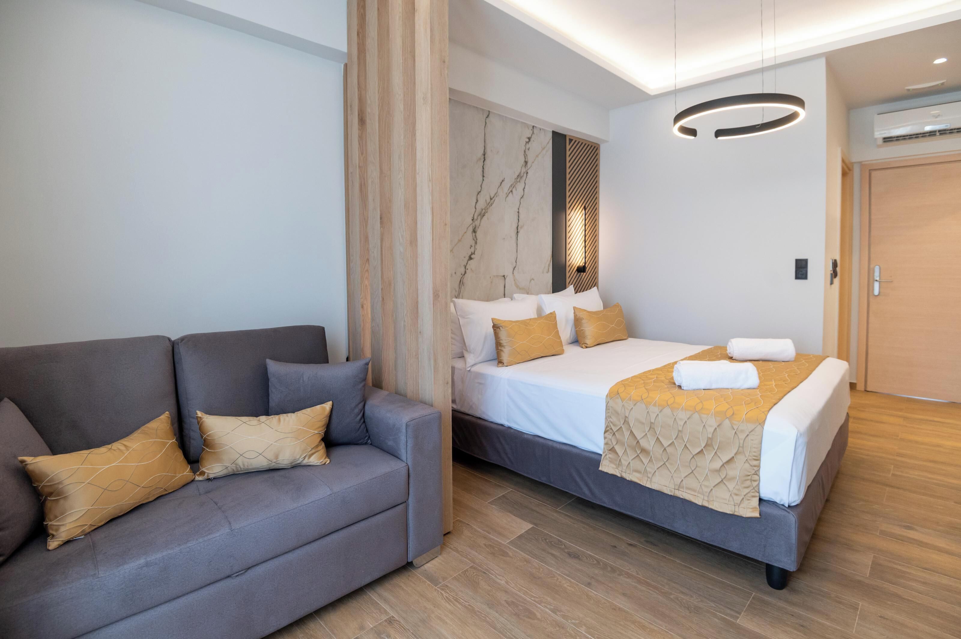 A modern hotel room with a grey sofa, a bed with a beige duvet, and a wooden floor.