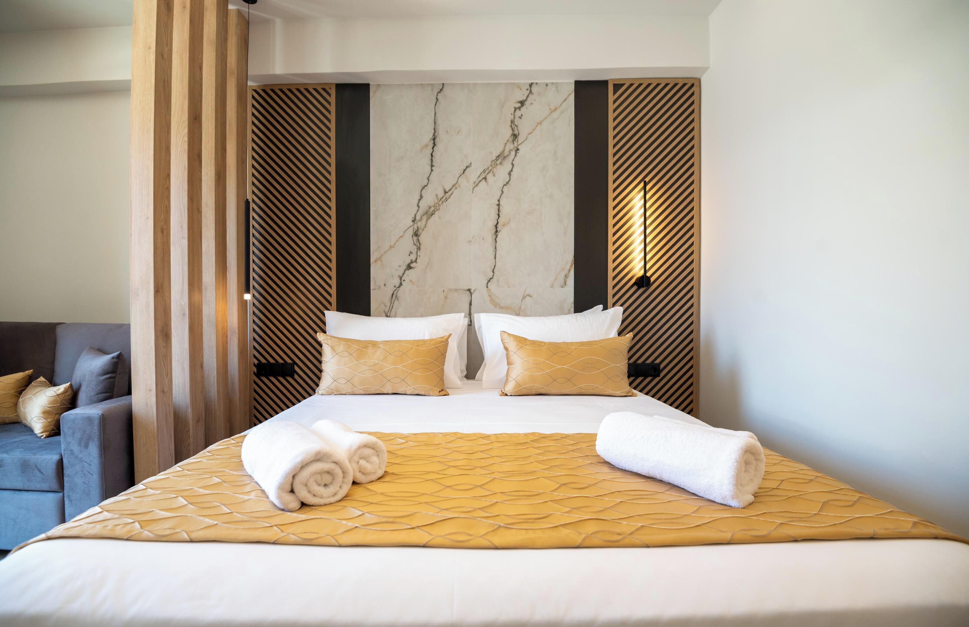 A hotel room with a large bed, two pillows, a rolled towel, a sofa, and a decorative wall panel.