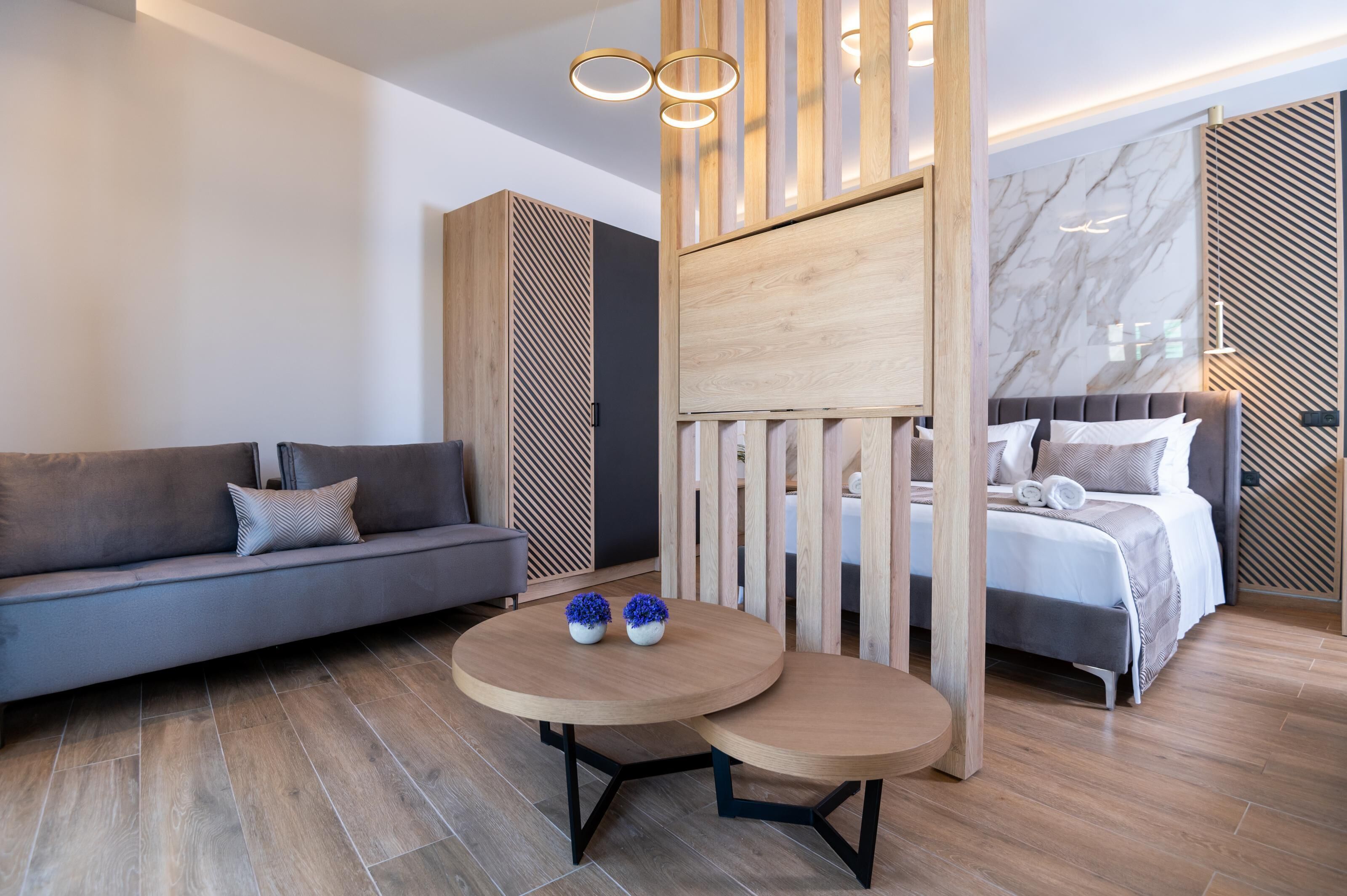A modern hotel room with a sofa, a bed, and a round wooden table with two vases.