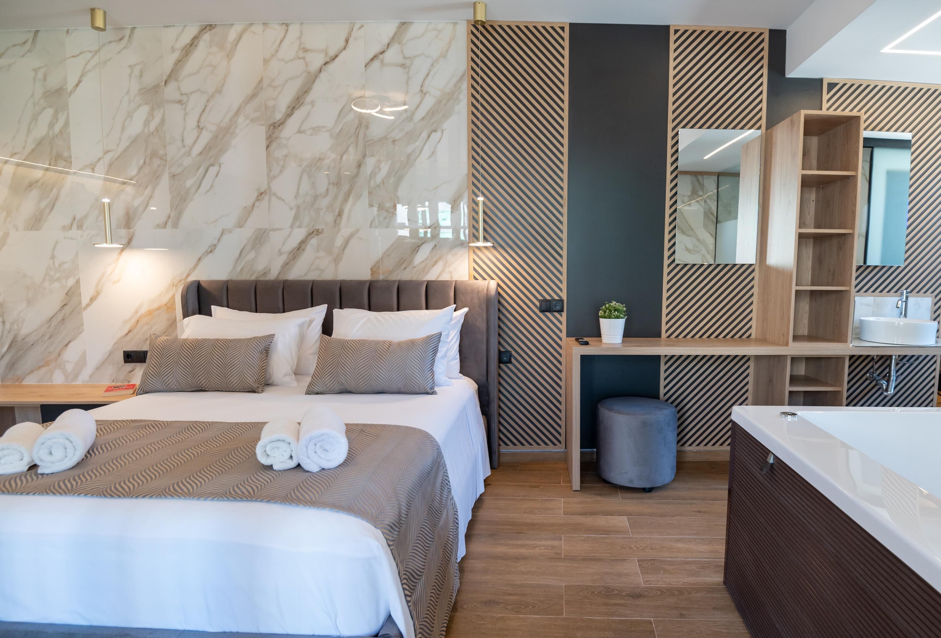 A modern hotel room with a marble wall, a large bed with a headboard, a wooden vanity with a mirror, and a separate bathroom with a bathtub.