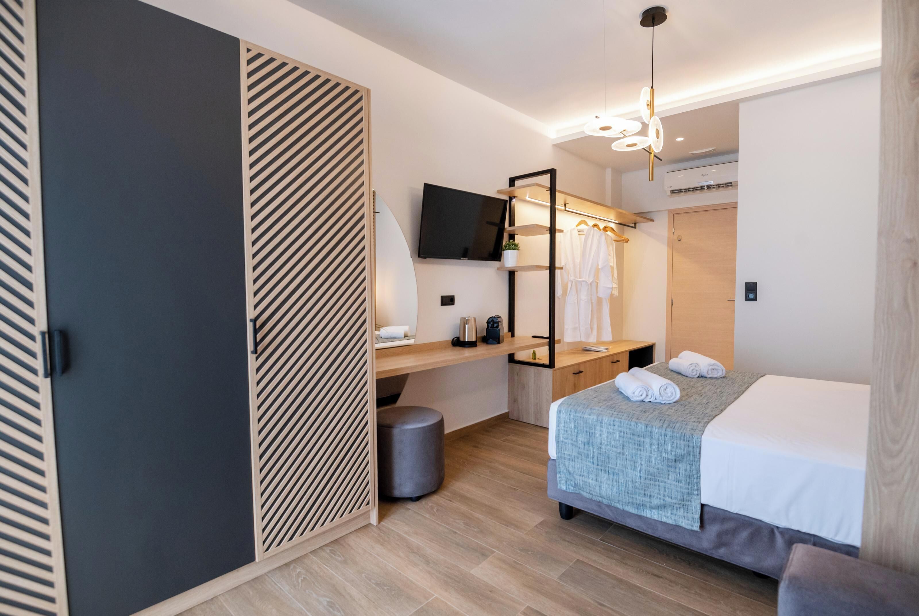 A modern hotel room with a bed, a desk with a TV, a wardrobe, and a hanging robe rack.