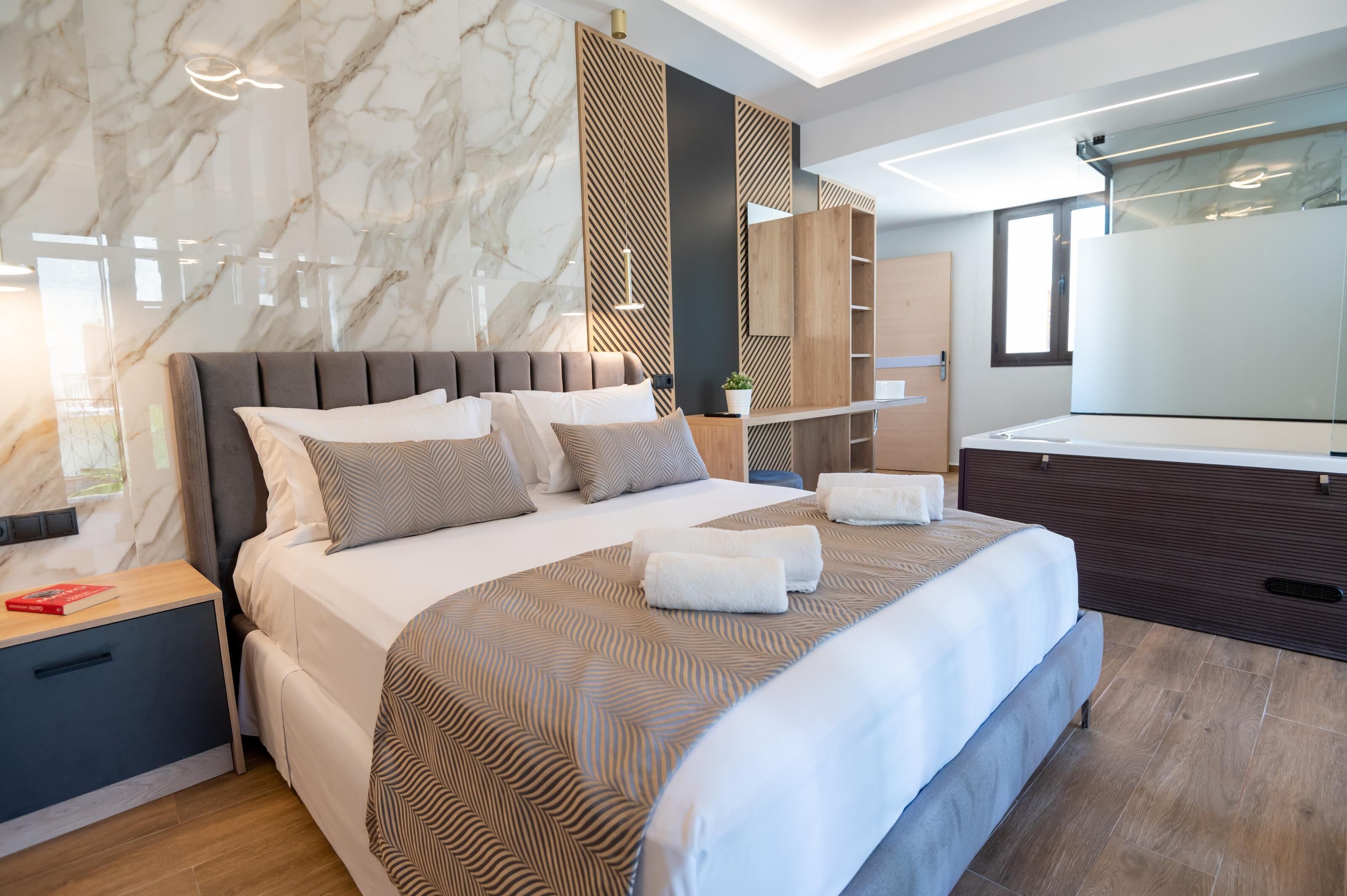Een moderne hotelkamer met een marmeren wand, een groot bed met een houten hoofdbord, een nachtkastje met een boek en een grote spiegel.