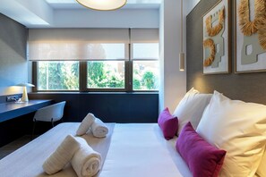 A modern hotel room with a large bed, a desk, and a chair. There are towels on the bed, and the room has a view of trees and buildings outside the window. - Feelathome Nervión Apartments (Seville)