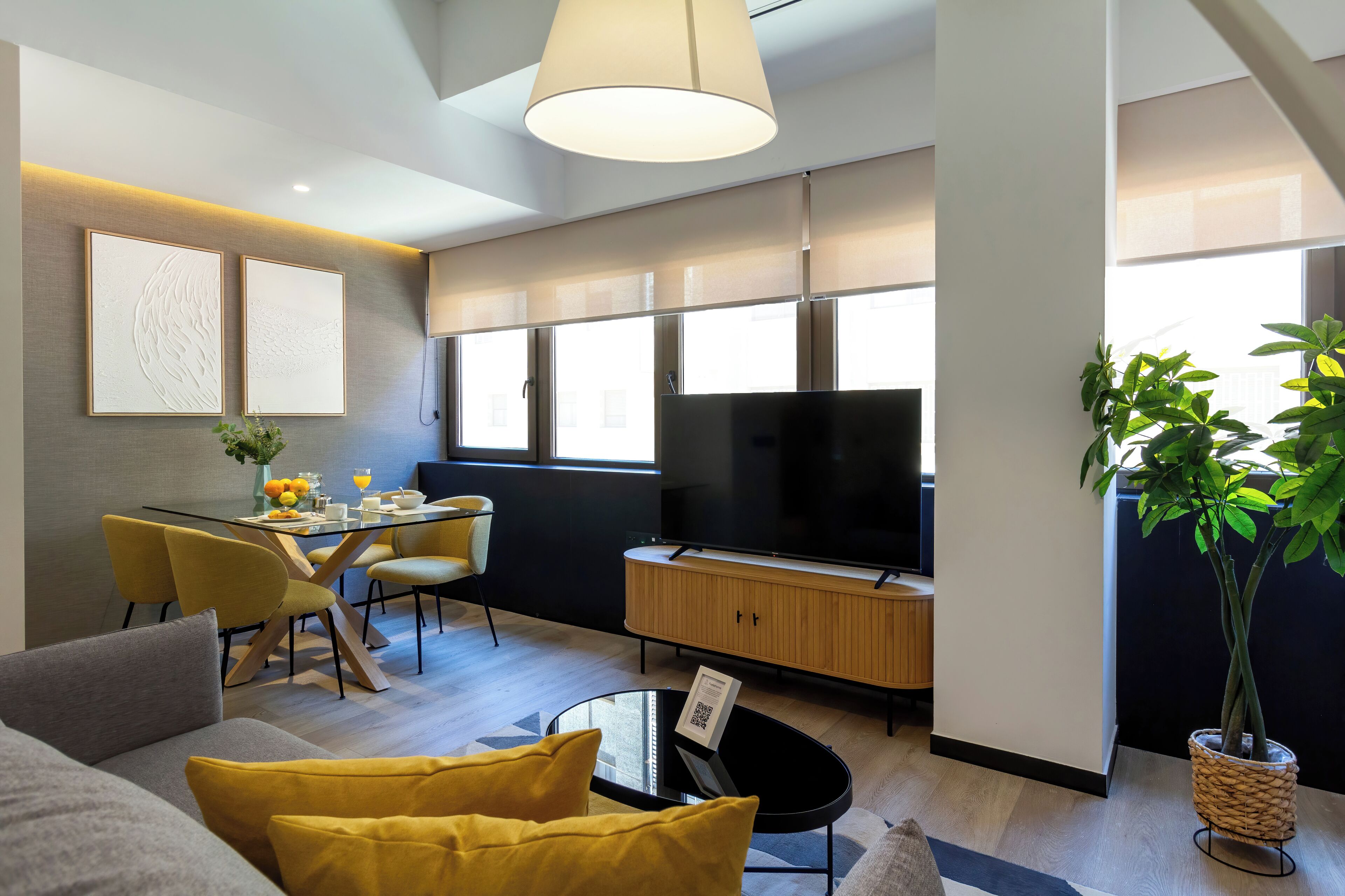 A modern living room with a dining area, a flat-screen TV, and a large potted plant.
