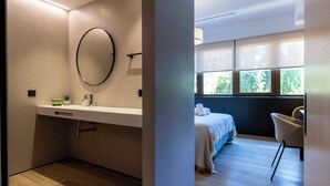 A modern bathroom with a large mirror, a white countertop, and a sink. There is a bed with a blue headboard and a chair in the adjacent room. - Feelathome Nervión Apartments (Seville)