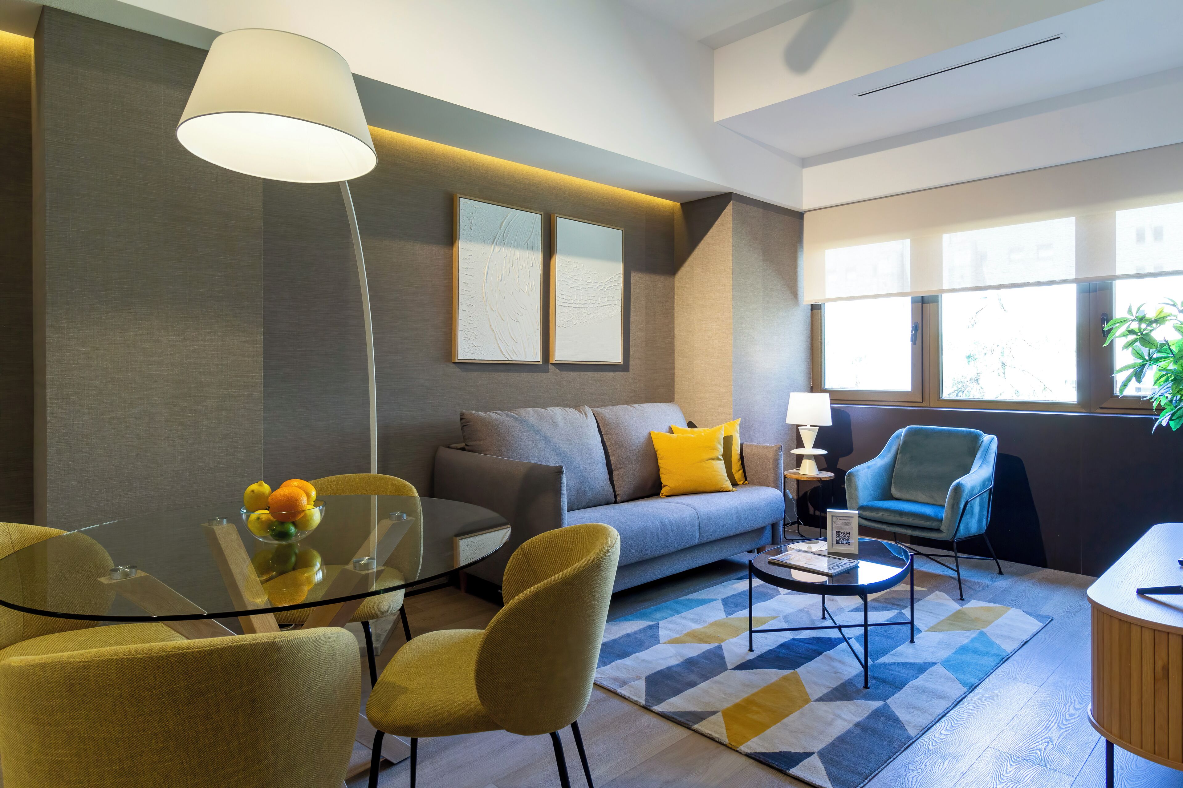 Una sala de estar moderna con un sofá, sillones amarillos, una mesa de vidrio con fruta y una lámpara.