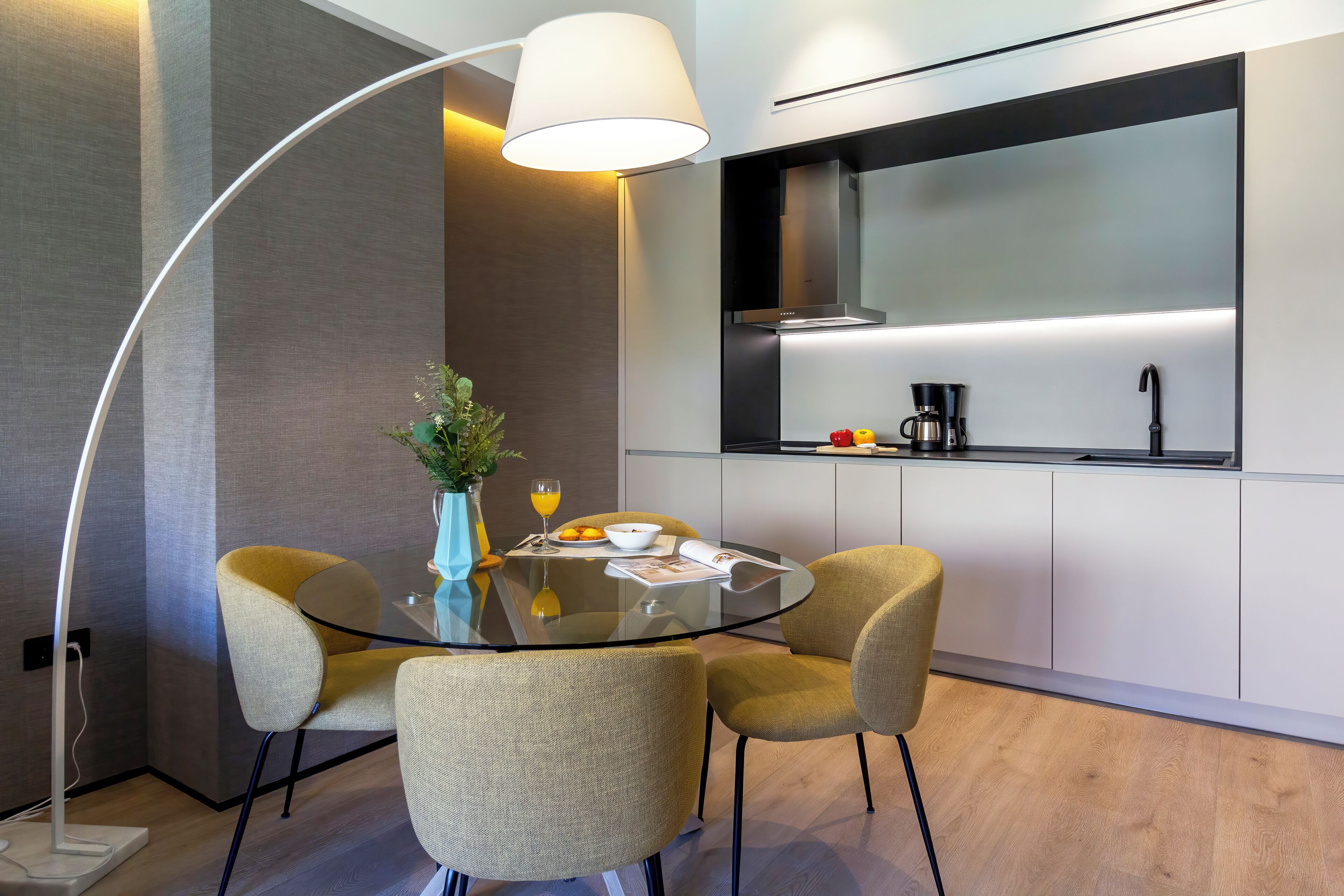 Een moderne keuken met een glazen eettafel, twee stoelen, een vaas met bloemen en een koffiezetapparaat.