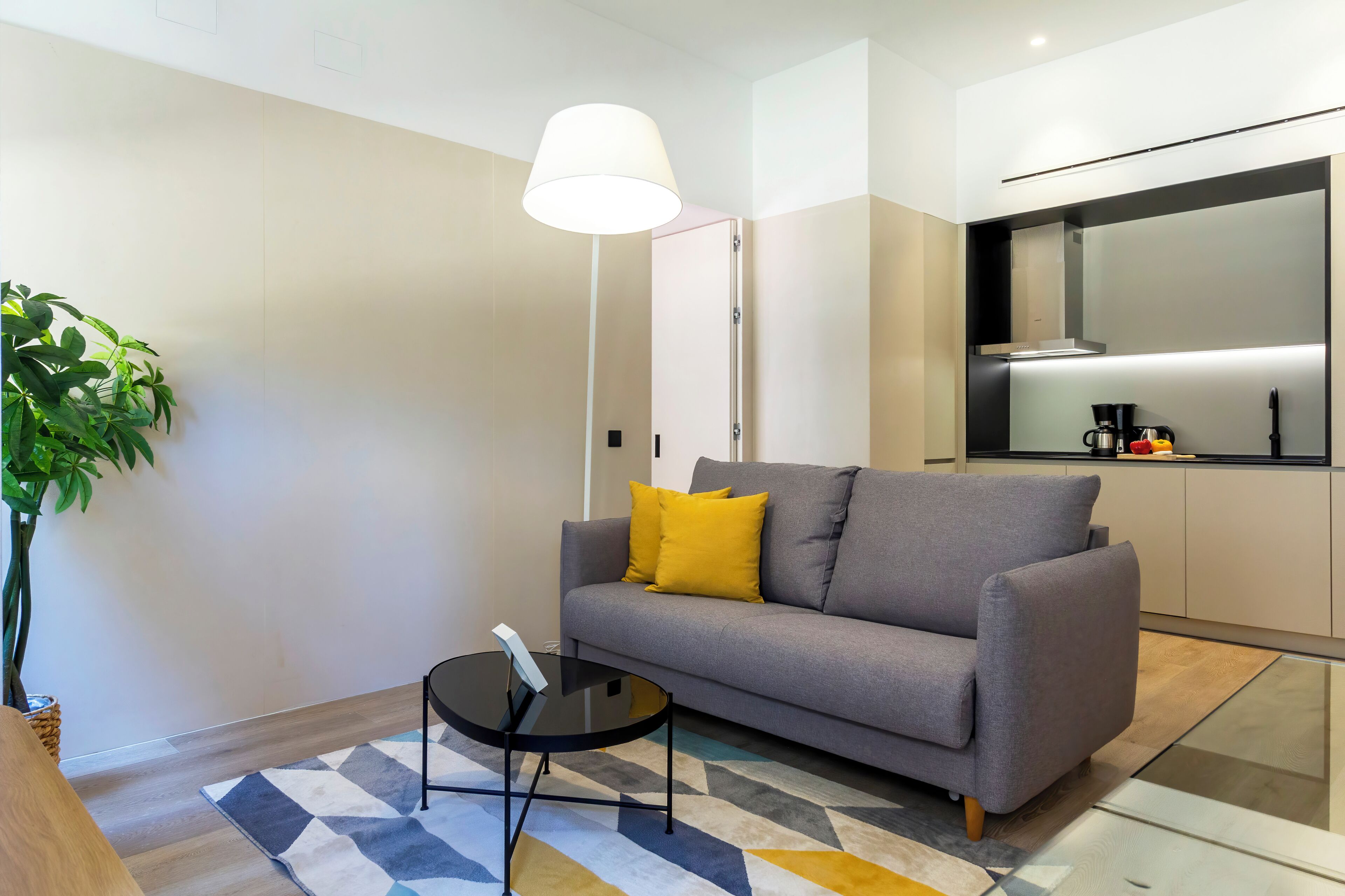A modern living room with a grey sofa, a small round table, and a floor lamp. A kitchen area is visible in the background with a coffee machine and a microwave.