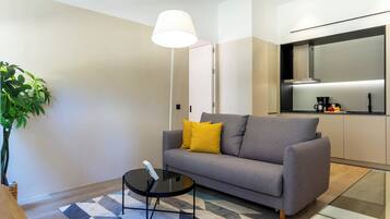 A modern living room with a grey sofa, a small round table, and a floor lamp. A kitchen area is visible in the background with a coffee machine and a microwave.