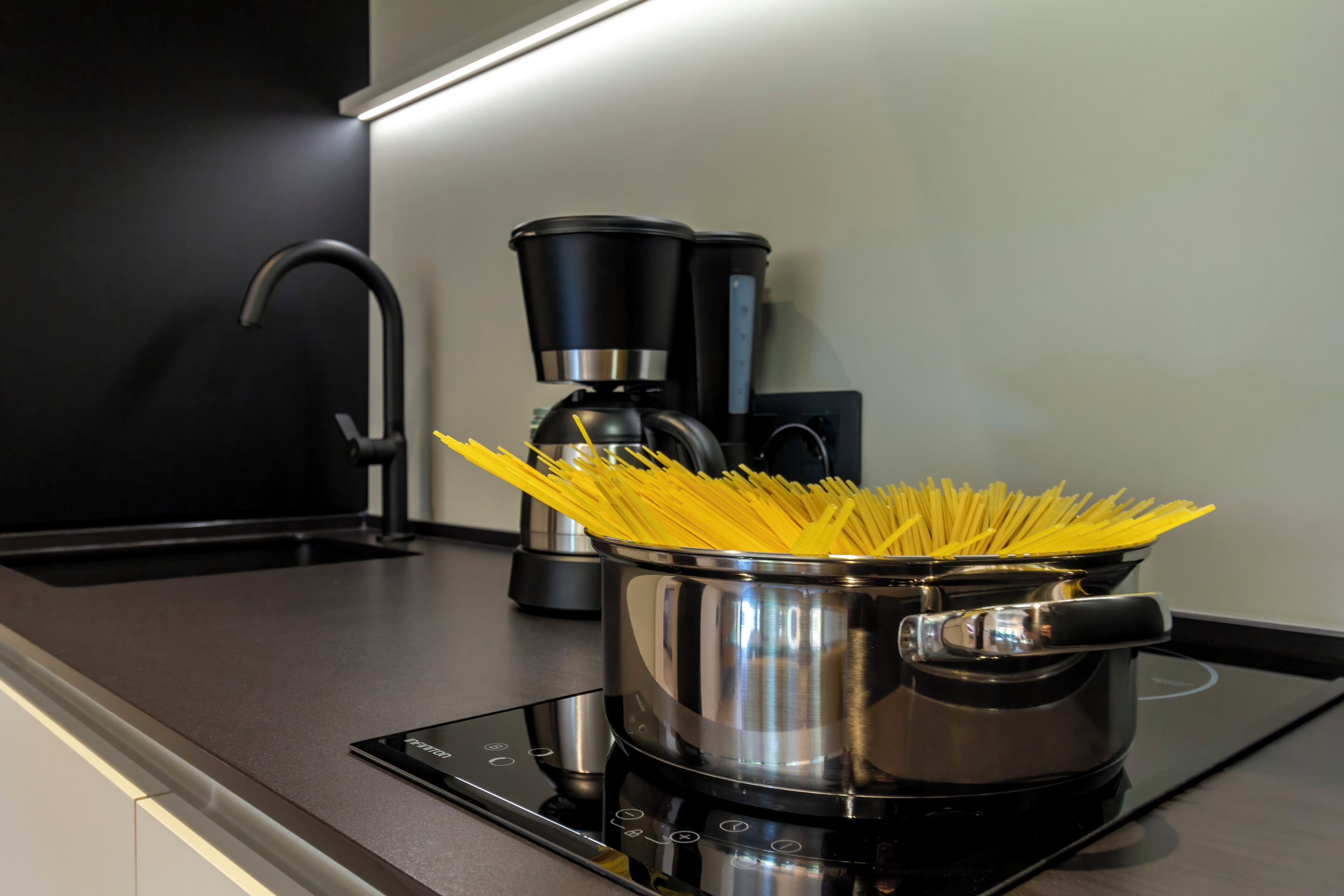 A kitchen with a pot of spaghetti on an induction cooktop, a coffee maker, and a modern faucet.