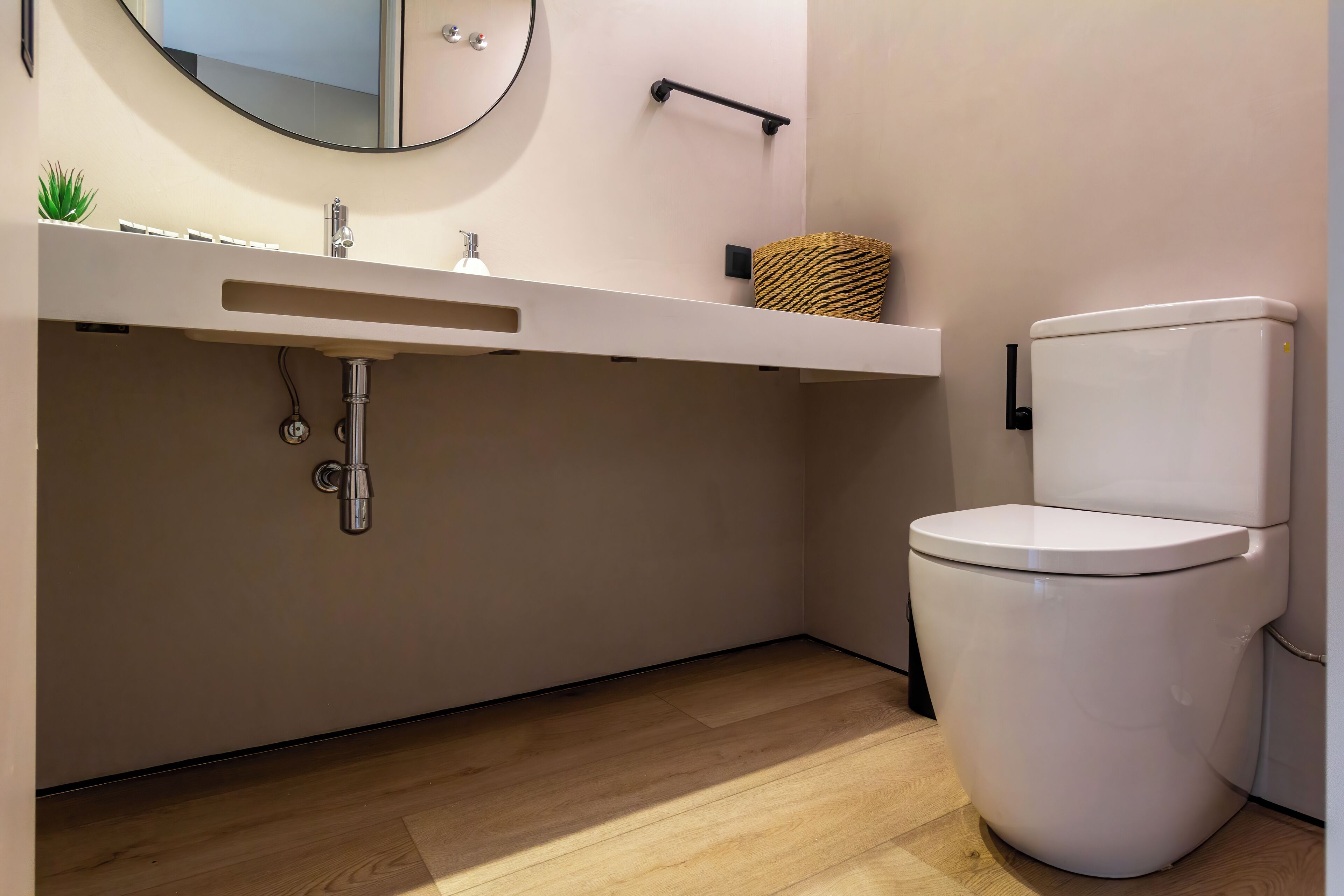 Un baño moderno con un lavamanos blanco, un espejo redondo, un inodoro y suelo de madera.