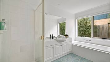 A modern bathroom with a glass shower enclosure, a white sink with a mirror above it, and a large window providing a view of greenery outside.