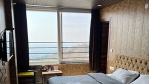 Habitación de hotel con una cama grande, una mesita con una tetera, una silla y vista al mar a través de un ventanal amplio.