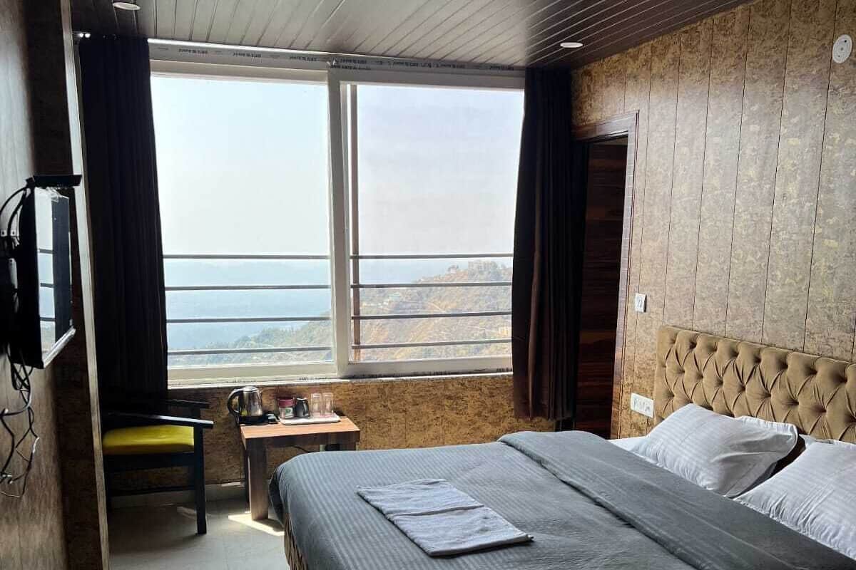 A hotel room with a large bed, a small table with a kettle, a chair, and a view of the sea through a large window.