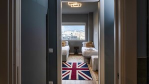 A narrow hallway leading to a bedroom with a view of a cityscape. The room has a wooden floor, a Union Jack rug, and two beds with pillows and a small bedside table. - Carignano Luxury Suite by Wonderful Italy (Genoa)