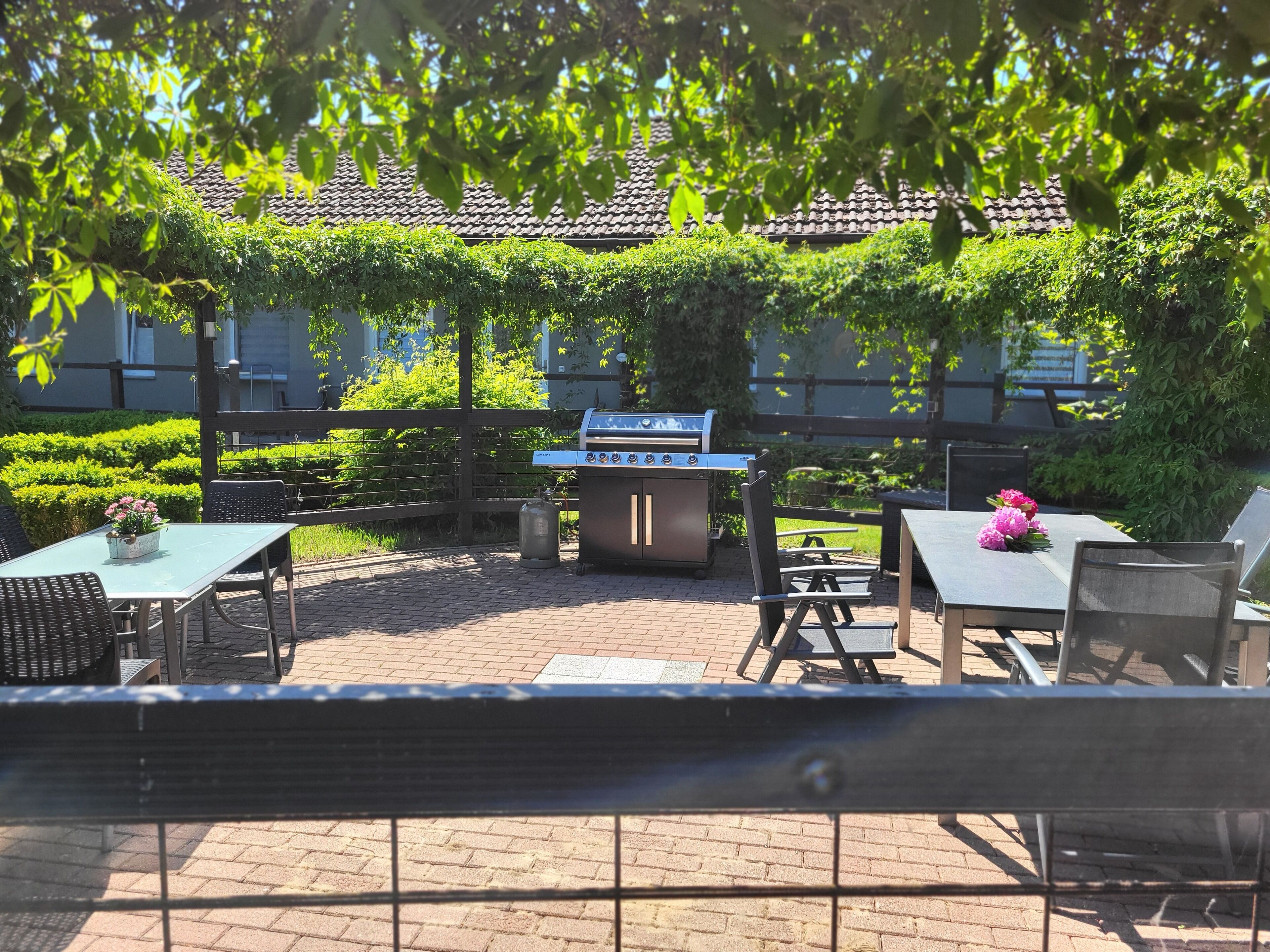 Patio esterno con barbecue, tavoli e sedie, circondato dal verde e una struttura a copertura in tegole.