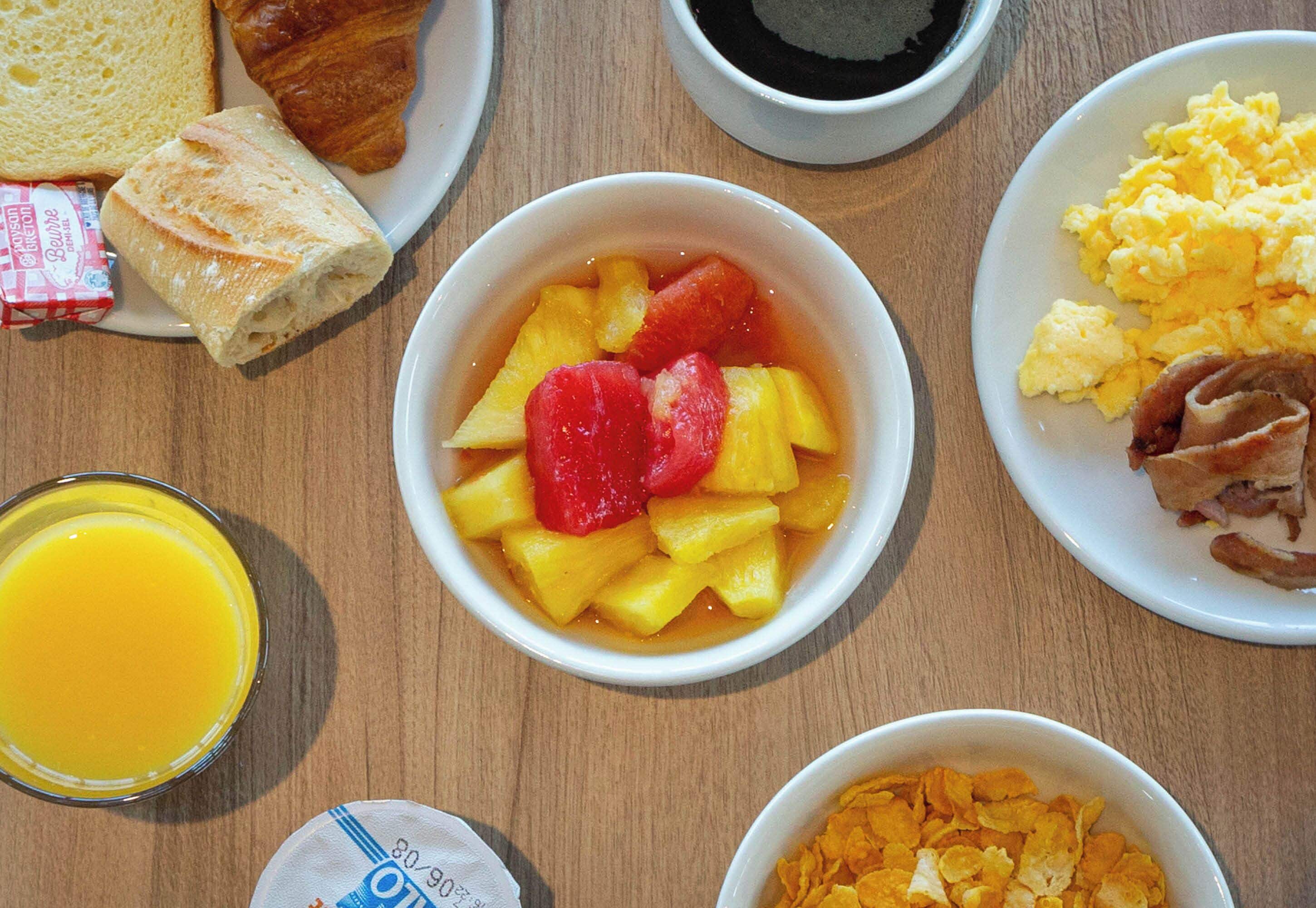 Een ontbijttafel met diverse soorten eten, waaronder toast, een croissant, een fruitschaal, roerei, spek, ontbijtgranen en een glas sinaasappelsap.