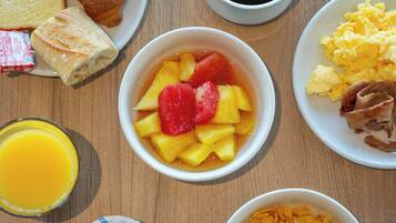 Een ontbijttafel met diverse soorten eten, waaronder toast, een croissant, een fruitschaal, roerei, spek, ontbijtgranen en een glas sinaasappelsap.