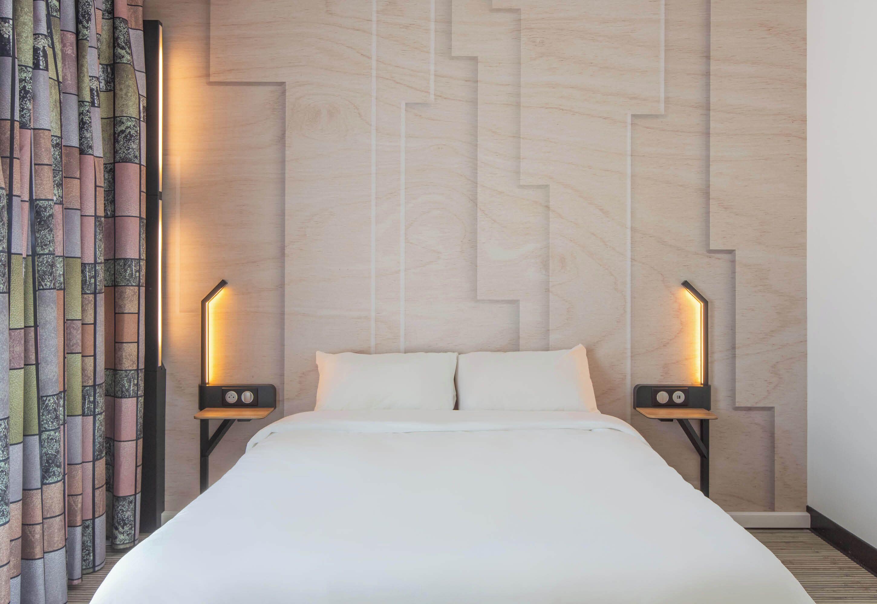 Habitación de hotel con una cama, dos mesitas de noche, una cortina estampada y una pared con un diseño geométrico.