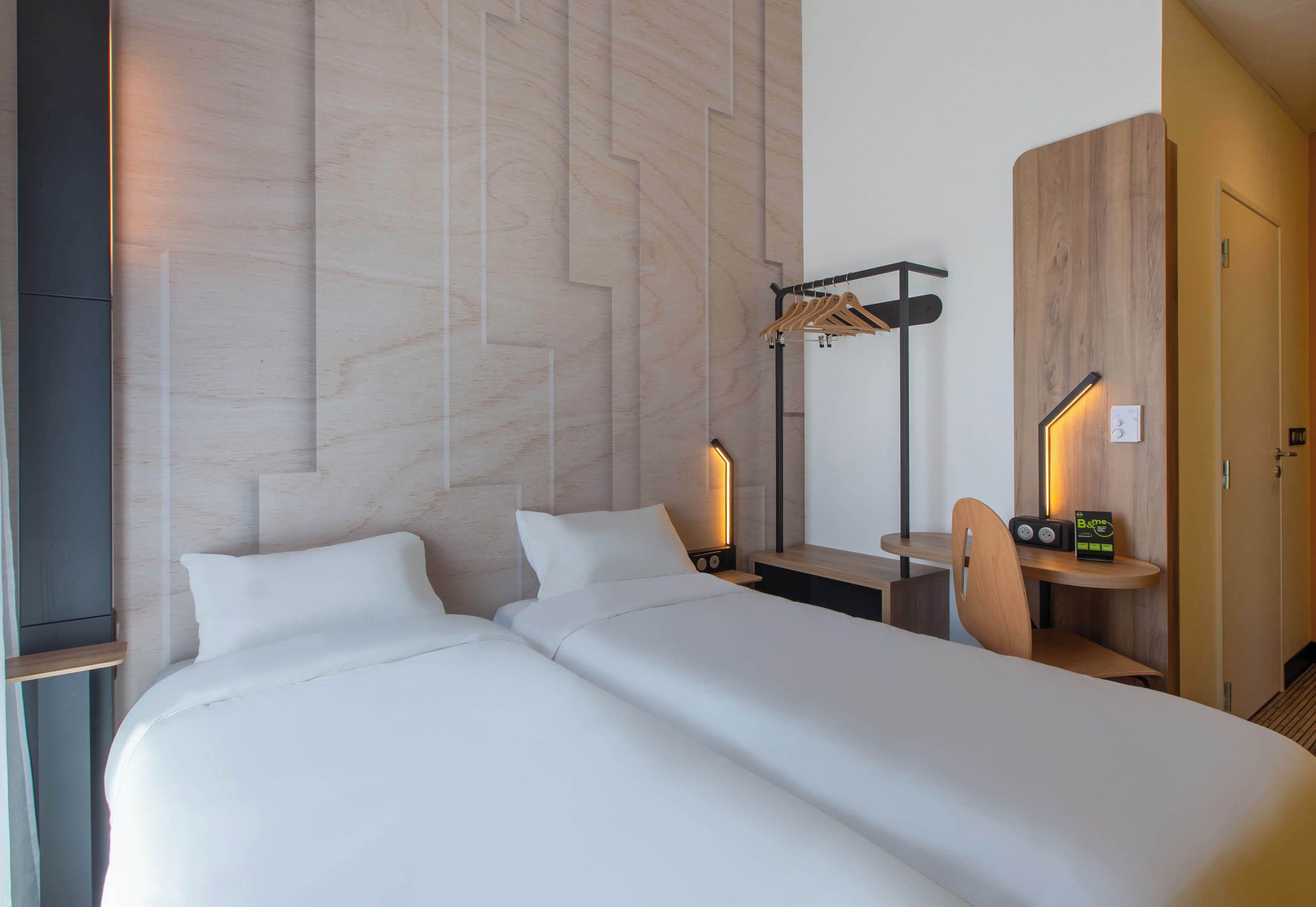 Habitación de hotel con dos camas, un escritorio, una silla, un perchero y una mesita de noche con lámpara.