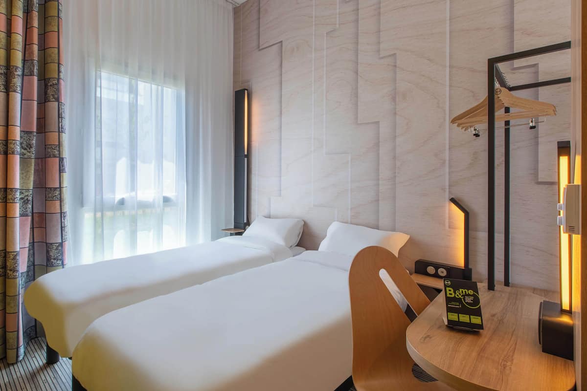 A hotel room with two beds, a wooden chair, a table with a B&Q sign, and a mirror with a wooden hanger.