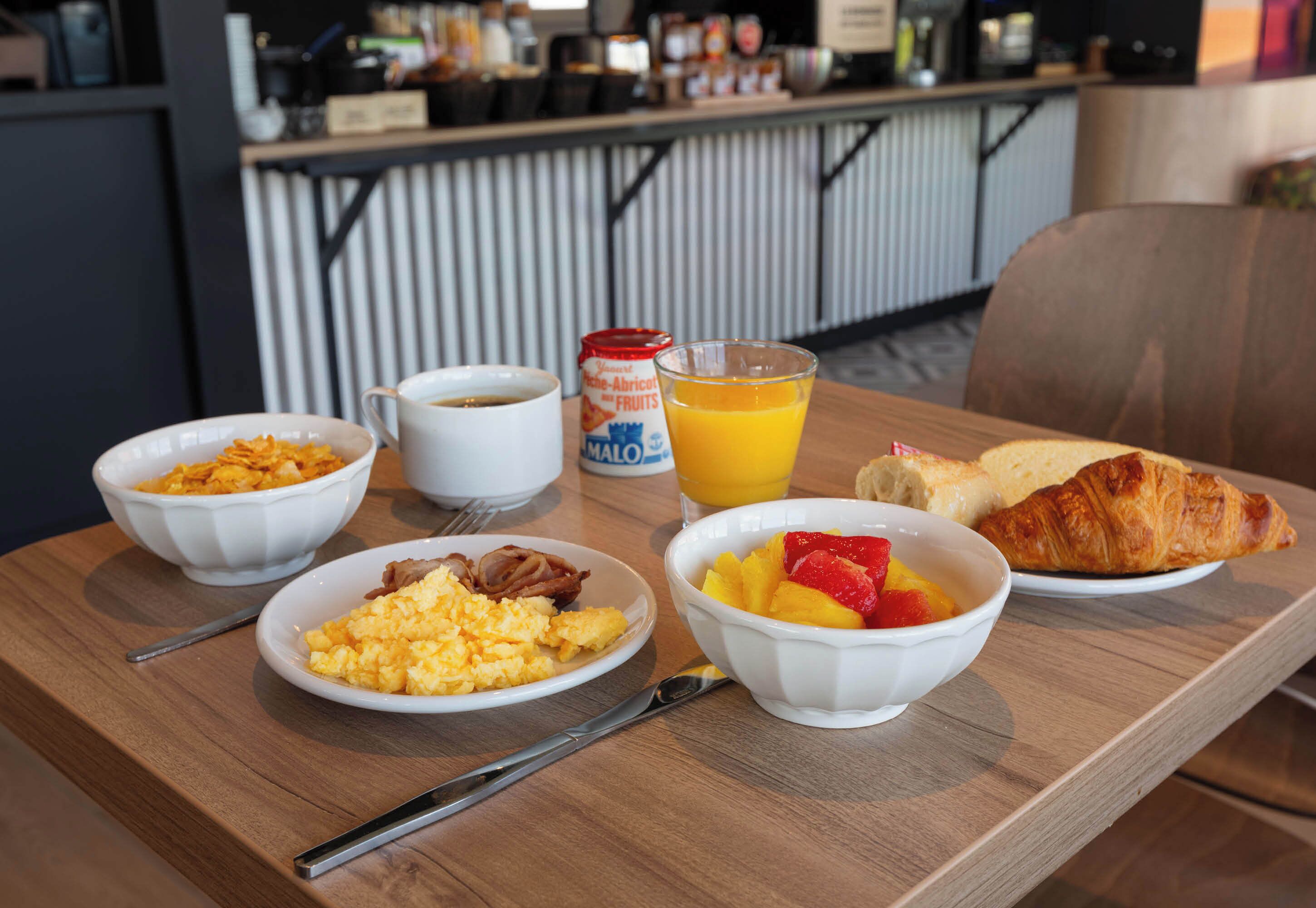 A breakfast table with scrambled eggs, bacon, fruit, orange juice, and a croissant.