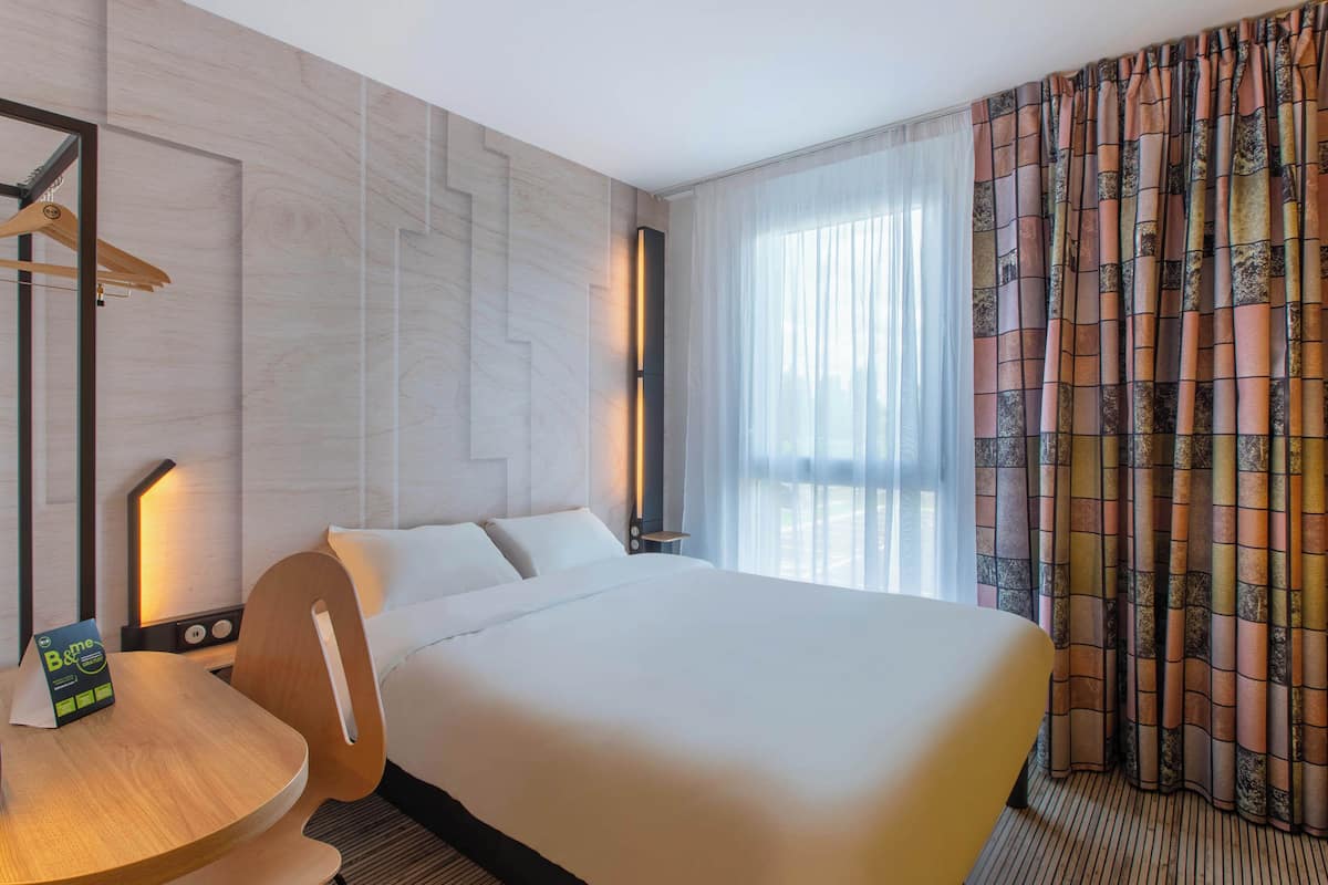 A hotel room with a large bed, a wooden chair, a small table, a nightstand, and a mirror.