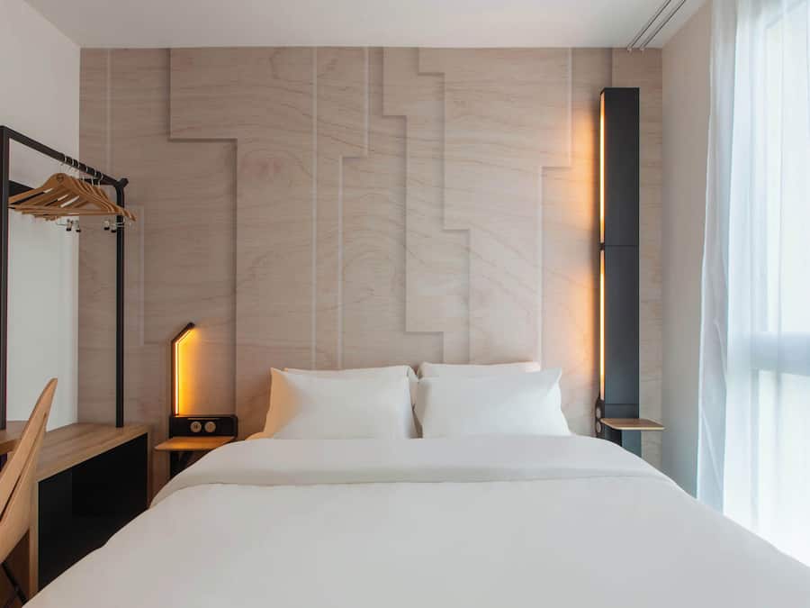 A modern hotel room with a large bed, a wooden desk, a nightstand with a lamp, and a wardrobe with a mirror.