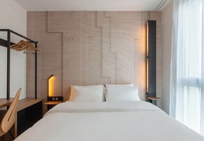 Una habitación de hotel moderna con una cama grande, un escritorio de madera, una mesita de noche con lámpara y un armario con espejo.