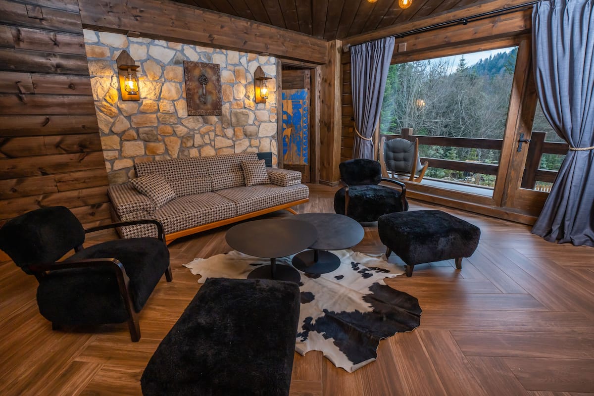 A log cabin interior with a stone wall, wooden furniture, and a large window with a view of trees.