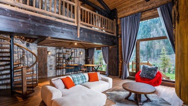 A spacious living area with a wooden staircase, a sofa with orange cushions, a red chair, and a large window offering a forest view. - Bi Abant Masali Village Hotel (Bolu)